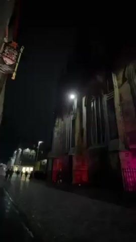 The video depicts a street scene at night. A large stone building, possibly a church or cathedral, is prominently featured on the left, with its base illuminated by red and pink lights. The street is paved with cobblestones, and a few people are walking along it. Streetlights provide additional illumination. The sky is dark, suggesting a late evening or nighttime setting. Other buildings line the street, with some displaying decorative lights.