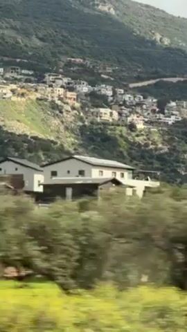 The video shows a hillside dotted with houses. The hillside is covered in green vegetation. The camera moves quickly, suggesting it is filming from a moving vehicle. A tall communication tower is seen near the road. The sky is cloudy.