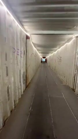This is a first-person perspective video of someone walking through a tunnel made of shipping containers. The tunnel is lit with white strip lights along the top on both sides. The walls of the containers are visible, showing some graffiti. The ground appears to be a dark paved surface with some white lines. The tunnel extends into the distance, with a brighter light visible at the far end.