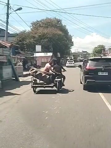 Two men are riding a motorcycle with a sidecar that is carrying large tires. The motorcycle is on a paved road with buildings and trees on either side. A black SUV is driving in the same direction as the motorcycle. Power lines are visible overhead. The sky is blue and the weather appears sunny.