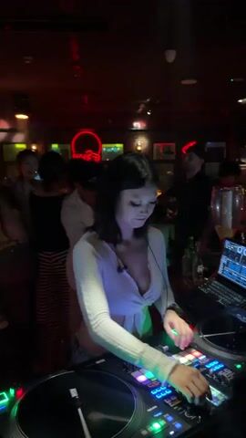 The video shows a young woman DJing in a dimly lit club or bar. She is wearing a white, crop top with a plunging neckline and has short, dark hair. She is focused on the DJ equipment in front of her, which includes turntables and a mixer with numerous colorful buttons and lights. A laptop is also visible on the DJ booth, displaying what appears to be DJ software.

In the background, other people can be seen, some dancing and others standing around. The lighting is atmospheric, with red and green neon lights creating a vibrant, energetic mood. Red neon signs in the shape of figures or objects are visible on the back wall. The overall impression is of a lively nightlife scene. The timestamp indicates the video is from 00:00 to 00:04, suggesting a short clip or the beginning of a performance. The location context provided is Liangjiang, China. No visible text is present in the video itself.