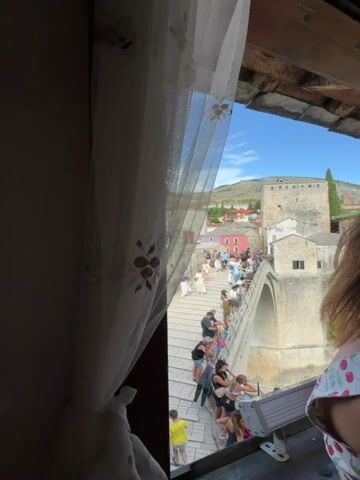 The video shows a view of the Stari Most bridge in Mostar, Bosnia and Herzegovina. The bridge is crowded with people, many of whom are tourists. Some people are walking across the bridge, while others are standing and looking at the view. There are also some people sitting on the edge of the bridge. The buildings on either side of the bridge are traditional stone buildings, and there are colorful houses in the background. The river Neretva flows beneath the bridge. The weather is sunny and the sky is blue. A curtain hangs in the foreground, partially obscuring the view. A person's shoulder and hair are visible in the bottom right corner of the frame.