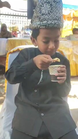 A young boy sits on a white chair, dressed in a black outfit and a patterned hat. He eats something from a small white cup with a red spoon.  There are blurred figures and a yellow draped table in the background. The scene appears to be outdoors, possibly at an event. A blue and white tent is visible in the background.