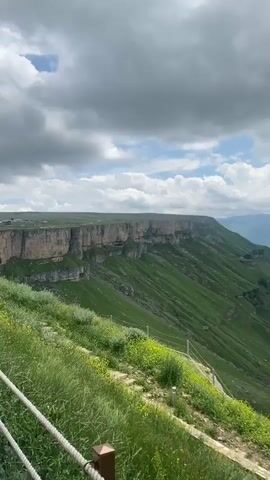 This video captures a scenic view of a vast, green landscape with a dramatic cliff face in the background under a cloudy sky. In the foreground, people are walking along a paved pathway next to a railing. A young girl in a denim jacket holds hands with an adult wearing a black and gold patterned dress. Another adult in a white headscarf and a dark outfit walks ahead. Further down the path, a group of tourists in casual attire are seen walking towards a building with large windows and a stone facade. The building appears to be a restaurant or cafe, with outdoor seating visible. The overall impression is a pleasant day out enjoying nature and sightseeing.