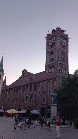 The video shows a city square in front of a large, brick building with a tall clock tower. People are walking around the square, some are sitting at outdoor tables, and there are streetlights and trees. In the foreground, there are several people on bicycles and scooters. The sky is a pale pink and purple, suggesting it is either dawn or dusk. The overall impression is one of a lively, urban environment.
