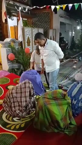 The video shows an outdoor evening gathering, possibly a celebration or religious event, with several people present. A man in a white shirt and blue jeans is standing and appears to be interacting with a group of people seated on the ground, who are covered with blankets or sarongs. Some of the people seated are children. Two young girls, one holding a white balloon, are also visible and interacting with an adult male. There are Indonesian flags displayed. The ground is covered with mats and rugs. Some decorations, including lights and banners, are visible. The overall atmosphere appears to be festive and communal.