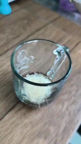 A clear glass mug sits on a wooden surface. Inside the mug, there is a small pile of light-colored granular material and a darker, powdery substance. Hot water is being poured from a silver container into the mug, over the materials. Steam is visible rising from the mug as the water mixes with the contents. The mug has a handle and faint markings on its surface. The background is blurred, showing hints of a white appliance and purple objects.