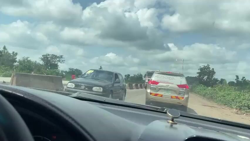 The video shows a view from inside a car, looking forward. The car is stopped in traffic on a road. Ahead of the car are several other vehicles, including a gold minivan and a large truck. The road appears to be under construction, with concrete barriers visible on the side of the road. The sky is cloudy.