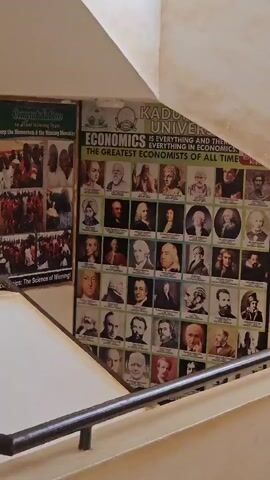 The video shows the interior of a building. A wall display showcases a poster titled "Kaduna University Economics" featuring pictures of notable economists. Adjacent to it, there's a second poster with the title "Congratulations to all our Winning Team".  The camera then pans to the right, revealing a hallway with a person walking away from the camera. The corridor is well-lit and features multiple doorways.