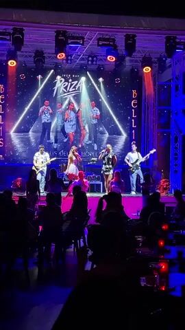 A live band named "PRIZA" performs on a stage under bright lights. The band consists of seven musicians: five singers in the background and three singers and two guitarists on the main stage. The audience is seated in a dimly lit area in front of the stage, facing the performers. The overall environment suggests a concert or performance venue, possibly a hotel or bar, with stage lighting and a backdrop displaying the band's name and hotel logo. The music appears to be upbeat, with the performers singing and playing instruments, and some audience members can be seen watching the performance.