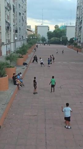 A group of young people are playing soccer in an open paved area between two tall apartment buildings. Several other people are sitting on a raised ledge to the left of the frame, watching the game. The sky is overcast.