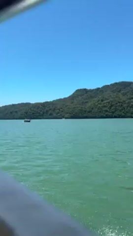 The video shows a view from a moving boat on a body of water under a clear blue sky. In the distance, there is a large, green, tree-covered landmass. Another boat is visible in the water, moving away from the camera. The water is a teal color and choppy from the movement of the boats.