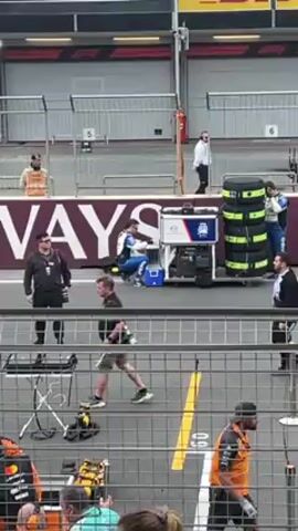 The scene is a view through a chain-link fence onto a Formula 1 pit lane area. Multiple individuals are visible. In the background, there's a large sign that reads "WAYS" in white letters. A figure in a blue and white racing suit is sitting on a small cooler next to a machine with screens. Other individuals dressed in race crew attire surround them, engaged in conversation or observation. To the right, there is a stack of tires wrapped in green tape. Other personnel in different attire, including one in a suit, walk across the pit lane. The environment suggests a race event, with pit lane markings and garage-style bays behind the scenes. A crowd is partially visible at the very bottom of the frame.
