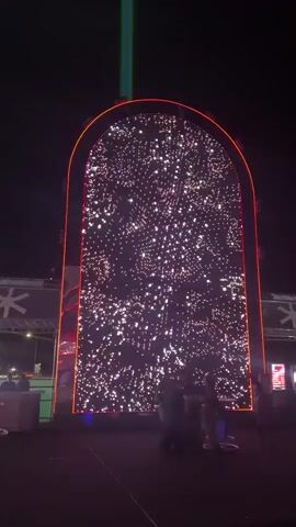 A large, arched LED screen displays a dynamic pattern of red and white lights that resemble sparkling stars or a digital nebula. The screen is framed by red lights around its perimeter. Below the screen, several people are visible, appearing to watch the display. The environment is dark, suggesting it is nighttime, and there are some structures or stage elements on either side of the screen, including what appear to be large snowflake graphics. The overall impression is of an outdoor evening event or exhibition featuring a light installation.