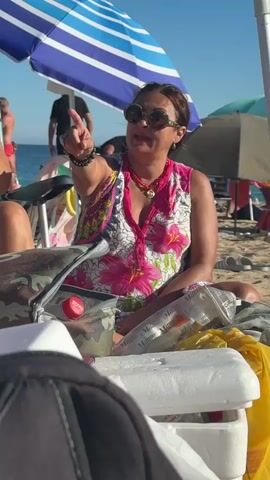 A woman is sitting on a beach under a blue and white striped umbrella. She is wearing sunglasses, a pink and white floral sleeveless top, and multiple necklaces. She has dark hair pulled back and is wearing bracelets on her wrists. She is gesturing with her left hand and speaking. In front of her is a cooler, a camouflage bag, and what appears to be bottles. Another person is partially visible on the left, wearing a bikini top and also wearing bracelets, with their left hand raised as if clapping or pointing. In the background, other people are visible on the beach, and the ocean is in the distance. The lighting suggests it is a sunny day.