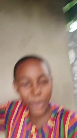 A young person with dark skin and short dark hair is smiling and looking at the camera. They are wearing a striped t-shirt with red, orange, yellow, green, blue, and purple stripes. Their right hand is raised and blurred, possibly waving or making a gesture. The background is a tan wall. The video is shaky and appears to be a selfie.