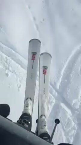 The video begins with a close up shot of skis on a ski lift, showing the skier's boots strapped in and ski poles at the bottom of the frame. The skis are light grey with red bindings and the word "AVION" on the tips. The camera then pans to show the ski slope below, covered in tracks and ski ruts. The shot continues to show the ski lift cable above a slope. In the distance, fog obscures the mountain scenery. The sky is overcast.