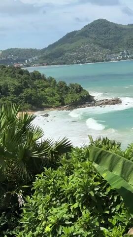 A high-angle, medium shot captures a scenic view of a tropical coastline. In the foreground, lush green foliage dominates, with large palm fronds and broad leaves obscuring the lower portion of the frame. Beyond the greenery, a turquoise ocean stretches out, with white waves crashing against a rocky shoreline and a small sandy cove. The water transitions to a deeper blue further out. In the middle ground, a verdant, tree-covered peninsula juts into the sea. In the background, a larger, mountainous island covered in dense green vegetation rises from the water. Along the slopes of this mountain, buildings and houses are visible, indicating a settlement. The sky above is partly cloudy, with patches of blue. The overall impression is a serene and beautiful coastal landscape.