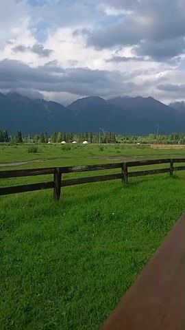 The video shows a scenic view of a grassy field with a wooden fence in the foreground. In the background, there are several white yurt-like structures and a line of trees. Beyond the trees, a mountain range is visible under a cloudy sky. The camera appears to be stationary, offering a static shot of the landscape. No people or animals are visible in the frame. The overall impression is of a peaceful, natural setting.