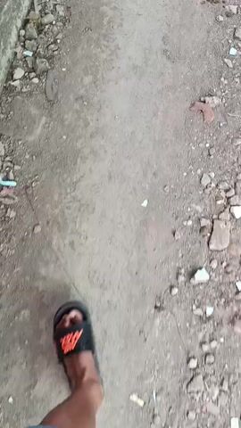 A person wearing black slides with orange lettering is walking on a path. The path appears to be made of dirt and gravel on the left side, with a sidewalk made of interlocking bricks on the right. There are pieces of debris scattered throughout the path, including what appears to be broken bricks, small pieces of plastic, and cigarette butts. The overall environment seems to be outdoors, possibly in an urban or industrial area given the debris and the rough texture of the path. The camera is focused on the person's feet and the ground, giving a first-person perspective of the walk. The lighting suggests it is daytime.