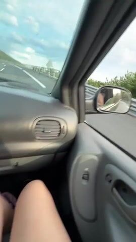 The video appears to be filmed from inside a vehicle, looking out the passenger side window. The perspective suggests the vehicle is in motion, driving on a road that runs alongside a bridge or elevated structure. In the foreground, the interior of the car is visible, including part of the dashboard and the side mirror. The side mirror reflects a person, likely the videographer or driver, wearing patterned clothing. The road itself is asphalt with a clear white line marking the edge. Beyond the road, there is a metal railing, characteristic of a bridge or overlook. In the distance, a hilly, tree-covered landscape is visible under a cloudy sky. The motion blur indicates movement, and the overall impression is a scenic drive.