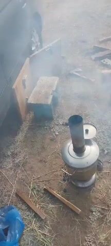 A video captures an outdoor scene with a samovar emitting smoke in the foreground. In the background, a wooden bench is positioned next to a wooden structure. Bricks are arranged on the ground, and scattered wooden debris is visible. The ground is covered with dry grass and dirt. The lighting suggests it is daytime. The camera is positioned overhead, looking down at the scene.