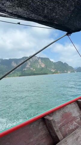 The video shows a view from the inside of a wooden boat moving through a body of water. The boat has weathered wooden planks and a red trim along the edge. A black mesh canopy is visible above, supported by metal poles and ropes. The water is a turquoise color, with ripples and wakes indicating movement. In the distance, a chain of lush green, mountainous islands rises from the water under a cloudy blue sky. The perspective suggests the boat is traveling towards these islands.