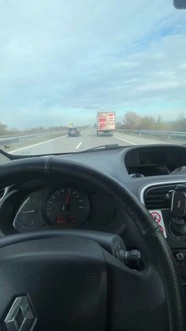 A static interior shot captures the driver's perspective from inside a car traveling on a highway during the day. The car's dashboard and steering wheel, black with a textured surface, dominate the foreground.  The speedometer needle hovers around 90 km/h. The windshield presents a view of a multi-lane highway stretching ahead. Other vehicles, including a large white truck with red branding, are visible in the distance. The sky is overcast with a mix of white and grey clouds. Trees line the sides of the highway. Accessories are visible on the dashboard, including a phone mount and a VIP-branded air freshener.