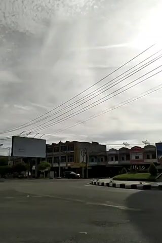 The camera films from a low angle, showcasing a prominent traffic circle featuring a large, wooden-looking sculpture in the shape of a betel nut. The sculpture is surrounded by manicured greenery and a sign that reads “SIGLI.” The surrounding buildings are visible in the background. The sky is overcast, but the sun still shines brightly. Power lines cross the frame above. A motorcycle and car pass by on the road.