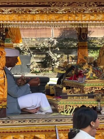 A man wearing a white head covering, a light grey jacket, and white pants sits on a raised platform, holding small items in his hands. He appears to be engaged in a ritual or ceremony. The platform is adorned with intricate gold and yellow fabric drapery and a decorative canopy. To his right, a golden dragon statue sits on a tiered altar, surrounded by offerings and decorative elements. In the foreground, partially visible, is a woman with dark hair, her back to the camera. The background shows a stone wall and lush green foliage, suggesting an outdoor or semi-outdoor setting, possibly a temple or cultural site. The lighting indicates it is daytime.
