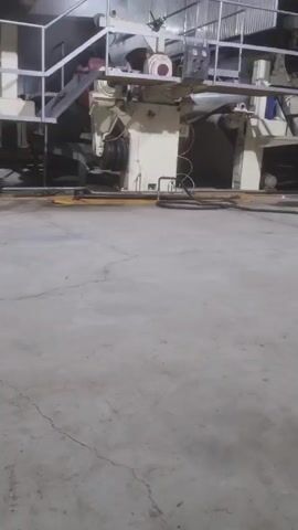 The video pans across a large, indoor industrial setting with a concrete floor, showcasing large machinery and metal structures. There are multiple levels, connected by stairs and platforms with railings. Two individuals, one in a hooded jacket, are seen walking in the background. A red fire extinguisher is visible near a concrete column. The environment appears dusty or foggy, adding to the industrial atmosphere.
