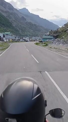 The video shows a point-of-view shot from a motorcycle or scooter on a paved mountain road. The road curves to the left and stretches ahead into a valley. On the left side of the road, there are several buildings with green roofs and some parked vehicles, including cars and what appear to be small trucks or buses. The right side of the road is lined with rocks and slopes upwards towards a mountainside with scattered buildings and trees. In the distance, large, rocky mountains rise up, with a cloudy sky overhead. A power line tower is visible on the right side of the frame. The perspective suggests forward motion along the road.