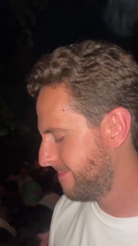 A man is recording himself in a crowded outdoor space with red and green lights. He has brown hair, a beard, and is wearing a white shirt. The camera focuses closely on his face, particularly his eyes, nose, and mouth. The background is blurry, but appears to be a party or concert atmosphere.