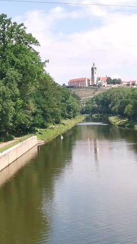 A wide shot captures a body of water flowing towards a distant town on a hill. The water is dark green and reflects the sky. On the left bank, a stone wall separates the water from a grassy area lined with trees. A white swan is visible on the grass near the water's edge. The town features several buildings, including a prominent church with a tall tower. Lush green trees cover the landscape on both sides of the water, and the sky is a light blue with scattered white clouds. The overall scene is peaceful and picturesque.