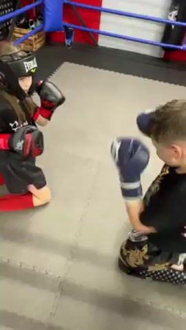 Two young children are sparring while kneeling, wearing protective boxing gear. The child on the left is wearing a black helmet with an Everlast logo, red and black boxing gloves, and black and red shorts. The child on the right has on dark blue and white boxing gloves and Thai-style shorts with gold designs. They are in a boxing ring, with blue, red, and white ropes visible in the background. The floor is covered in grey, square mats.
