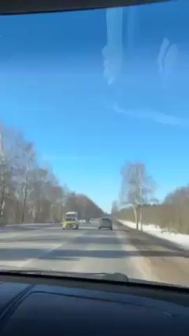 A car travels on a two-lane road with trees on both sides, snow on the shoulder, and a clear blue sky above. Several other vehicles, including a taxi and a van, are on the road. The view is from inside a car, showing the dashboard.