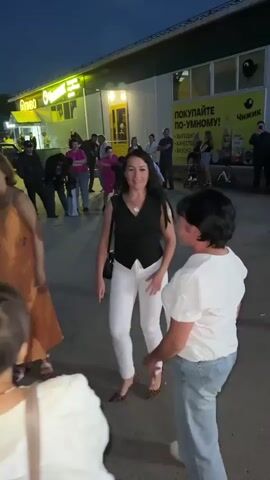 The video shows an outdoor scene at dusk in front of a building with a yellow awning and signs. Several people are present, some of whom are dancing. In the foreground, three women are the primary focus. The woman on the left wears an orange dress and is dancing with her arms outstretched. In the center, a woman in a black vest and white pants is also dancing. To the right, a woman in a white shirt and blue jeans is facing the dancer in the center and appears to be interacting with her, possibly also dancing or observing. In the background, a crowd of people watches and some appear to be filming. The environment suggests a public gathering or event.