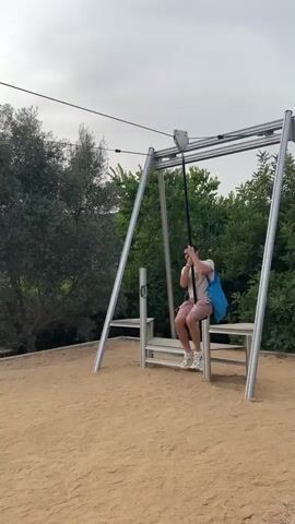 A man is using a zipline in an outdoor park. He is wearing a tan t-shirt, brown shorts, and white sneakers. He has a blue bag slung over his shoulder. The zipline has a grey pulley system attached to a wire, and the man is holding onto a black pole that is attached to the pulley. He is seated on a black seat. The ground is covered in sand. There are trees and buildings in the background. The sky is overcast with the sun peeking through.
