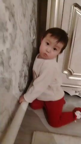 Here's a description of the video:

A toddler is kneeling on the floor, leaning against a wall. The wall is covered in wallpaper with a floral pattern. The toddler is wearing a white long-sleeved shirt with a smiley face and red leggings. They have dark hair.

The toddler looks up at the camera, makes a surprised or questioning face, and then smiles, reaching out with their hand.  A piece of furniture, like a cabinet, with a silver framed door is visible to the right. A white rug is on the floor.