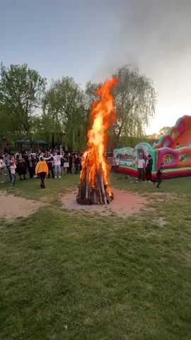 The video shows a large bonfire burning in a grassy area with a crowd of people standing around it. There is a colorful inflatable structure nearby. The people are standing and some are raising their arms. There are trees in the background and the sky is a light blue.