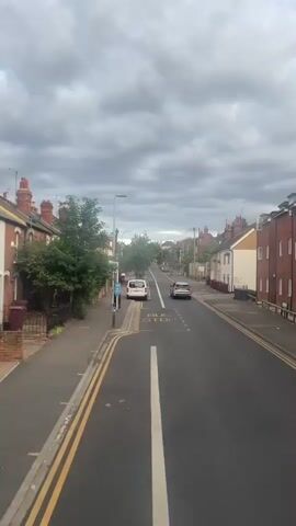 The video captures a street view from a moving vehicle, likely a bus or car, on a cloudy day. The road is paved and has distinct lane markings. Yellow double lines mark the edge of the road on the left side. On the right side of the road, there is a bus stop clearly marked with painted text on the asphalt. Several cars are visible on the road, some parked and others moving. Buildings line both sides of the street, appearing to be residential homes with brick and painted facades. Green trees and bushes are present along the sidewalk and in front of some houses. The sky is overcast with dense clouds. The camera is angled slightly downwards, looking ahead along the street.