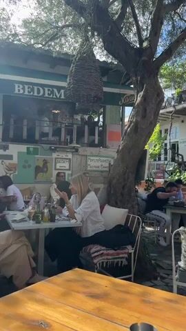 The video captures a daytime scene in an outdoor restaurant setting. Several people are seated at tables, engaged in eating and conversation. Some individuals are looking at their phones. Above the tables are trees that provide shade, and woven objects are hanging from the trees. The restaurant has outdoor art installations and signs visible. The sky is blue.
