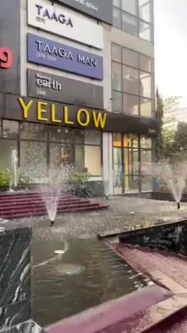 The video depicts the exterior of a building complex featuring shops and a small water fountain feature in front. The building has signs for "Taaga," "Taaga Man," and "Yellow," indicating retail businesses. The fountain feature includes several small, geyser-like water fountains spraying water into a shallow pool with angled edges. The foreground and background show some greenery and trees, while power lines are visible overhead. The video appears to be taken on a cloudy day.