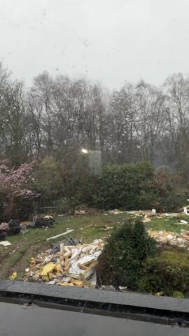 The video captures an outdoor scene, likely a backyard or an undeveloped plot of land, viewed through a window. The weather is overcast, and light snow or rain is falling, visible as numerous small white specks on the window glass and in the air, creating a soft, blurred effect in the background.

In the foreground, a dark window frame or sill is visible at the bottom of the screen. Beyond the window, the ground is covered in green grass interspersed with significant amounts of debris. This debris includes piles of white and yellow insulation material, pieces of wood, black plastic sheeting or bags, and a central mound of what appears to be burnt material. Several dark, possibly black, bins or containers are also scattered among the clutter.

To the left, there are dense green bushes and a tree with delicate pink blossoms, suggesting it might be a cherry or similar flowering tree, adding a touch of color to the otherwise muted palette. As the camera slowly pans to the right, more of the messy yard comes into view. A yellow garden hose snakes across the grass from the right side of the frame towards the center. On the far right, a small, conical evergreen tree stands amidst the debris.

In the mid-ground and background, there are numerous trees. Many are bare, indicating winter or early spring, while some are evergreen bushes or trees. A faint light source, possibly a street lamp, can be seen through the bare branches in the distance. The overall impression is one of a somewhat disheveled and untended outdoor area, possibly undergoing renovation or cleanup, under gloomy weather conditions.