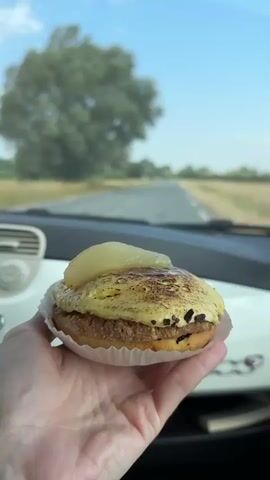 A person's hand holds a pastry topped with a slice of pear and caramelized cream. In the background, a road stretches through a rural landscape with fields and trees under a clear blue sky. The video appears to be filmed from inside a car.