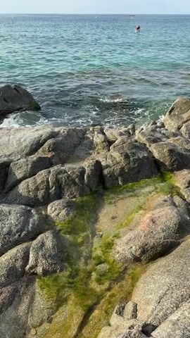 A medium shot captures a rocky shoreline with clear blue water gently lapping at the rocks. In the distance, a person is visible in the water, appearing to be on a paddleboard or similar floating device. The sky is clear with a few wispy clouds. The focus is on the interplay of the rocks, water, and the distant figure, creating a serene coastal scene.