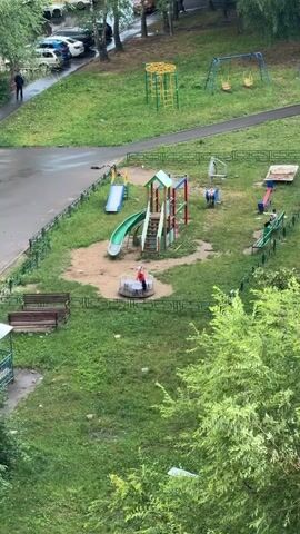 This media file captures an aerial view of a playground in Kolomna, Russia. The playground is equipped with a slide, swings, a merry-go-round, and a sandbox. A person in a red jacket is visible on the merry-go-round, while two other individuals are seated in a gazebo in the lower left portion of the frame. Cars are parked on a wet asphalt path bordering the playground, suggesting it may have recently rained or is currently raining. The presence of green trees and grass indicates it is likely daytime, possibly late afternoon or early evening. The overall scene depicts a quiet, everyday moment in a residential area.