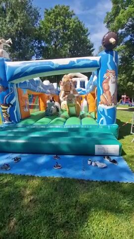 A woman walks onto a blue and green bounce house decorated with ice age themed characters. The bounce house is on a grassy area surrounded by trees. There are several pairs of shoes on the blue mat in front of the bounce house.
