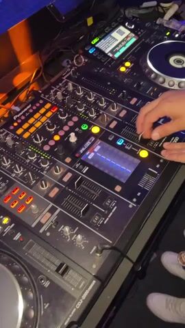 A close-up shot shows a person's hands operating a Pioneer DJ mixer and two CDJs. The mixer is black with numerous knobs, buttons, and sliders, many of which are illuminated with orange and green lights. A small screen on the mixer displays a waveform. The CDJs are also black, with large jog wheels and screens. One of the person's hands is on a knob on the mixer, while the other is resting on the mixer. The background is dark and appears to be a club or performance space, with a purple light visible on the top left. The bottom right of the frame shows the legs of two people wearing white sneakers. The timestamp at the bottom of the frame indicates the video is 5 seconds long.