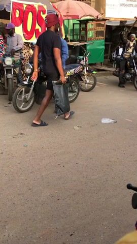 The video opens with a close-up shot of a motorcycle. The camera pans slightly to the right to reveal a man in a blue shirt and a dark blue cap sitting on a parked motorcycle. He is looking towards his left. To his right, another man in a black t-shirt and a red cap is walking away from the camera, carrying a black plastic bag in his right hand. He is wearing black shorts and flip-flops. In the background, there are several more motorcycles parked, some of which are occupied by people. There are also market stalls visible, with awnings and signs, one of which appears to say "POS". The ground is paved with asphalt, and there is some litter scattered on the ground. The scene appears to be a busy street or marketplace. The camera then pans again to the right, capturing more of the market activity and people. A person on a red motorcycle is seen approaching from the right side of the frame. The video continues to show snippets of activity in the market, with people moving around and motorcycles coming and going. The lighting suggests it is daytime, possibly late afternoon given the shadows.
